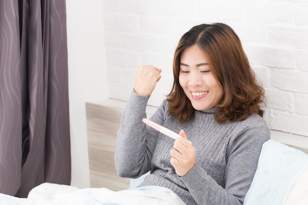 Happy asian woman holding pregnancy test and lying down in the bed