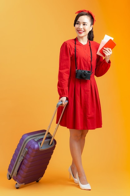 オレンジ色の背景で隔離の航空券とスーツケースとパスポートを保持している幸せなアジアの女性