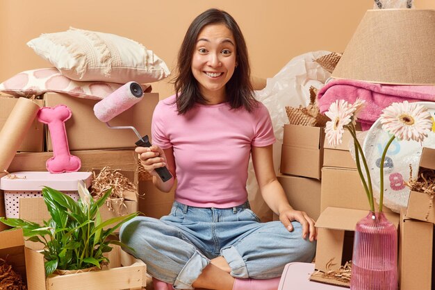 Happy asian woman holds paint roller to improve apartment\
relocates to new flat surrounded by cardboard boxes full of\
household items against beige background people real estate and\
mortgage concept