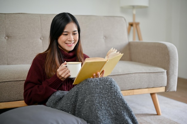 幸せなアジア人女性がホットチョコレートを飲みながらリビングで本を読んでいる