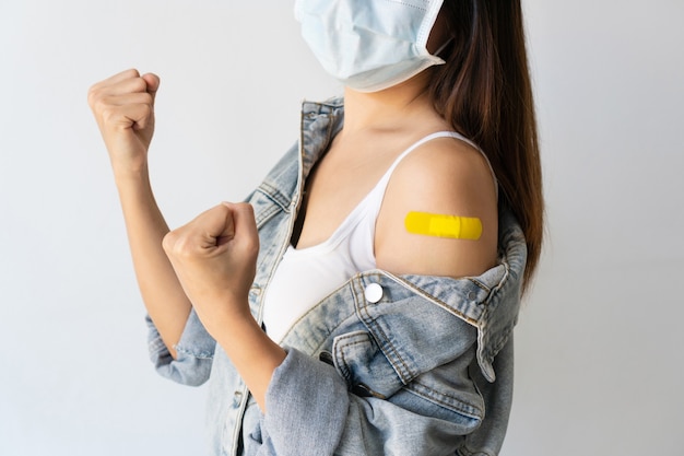 счастливая азиатская женщина в медицинской маске показывает руку с лейкопластырем после инъекции вакцины covid-19