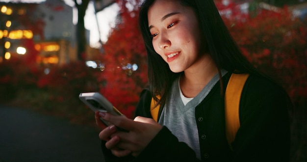 幸せなアジア人女性 電話と夜の街でソーシャルメディアコミュニケーションやアウトドアネットワークのために 女性の人が遅く夕方に携帯スマートフォンで笑顔でオンラインチャットする 日本の都市部の町