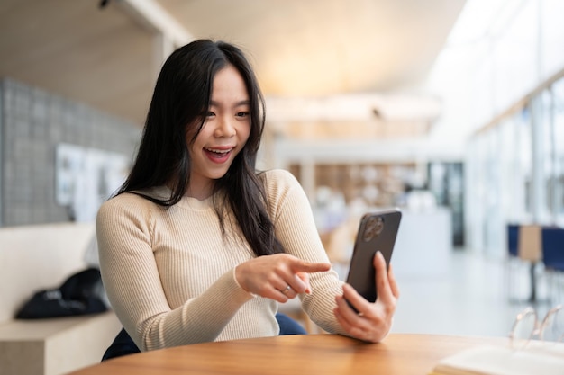 A happy Asian woman pointing her finger at her smartphone screen while sitting at a table