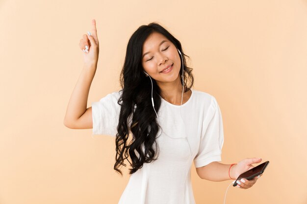 Happy asian woman posing isolated over beige wall space listening music with earphones by phone.