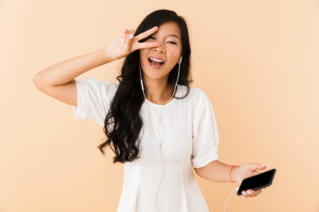 Happy asian woman posing isolated over beige wall space listening music with earphones by phone.