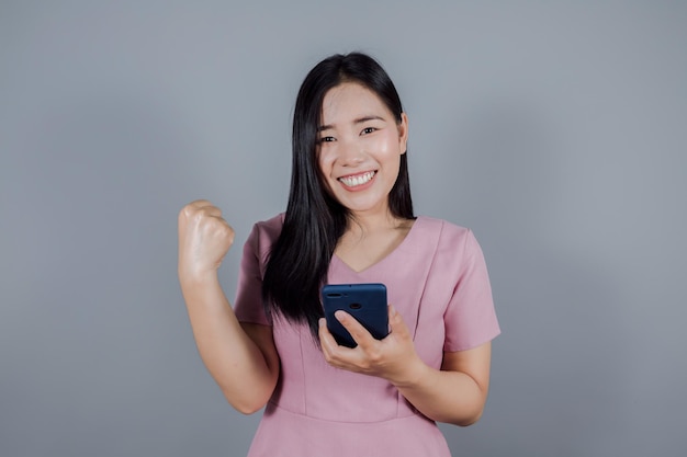 Счастливая азиатка поднимает руки, рада, взволнована, весела и держит мобильный телефон или смартфон на сером фоне
