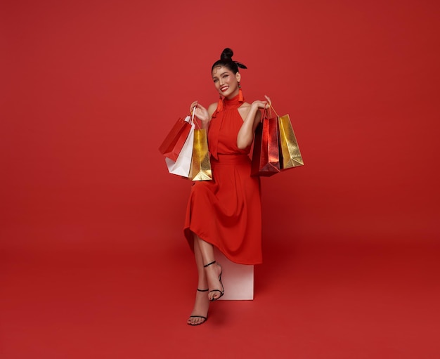 Happy asian woman in red dress holding shopping bags sitting on white chair