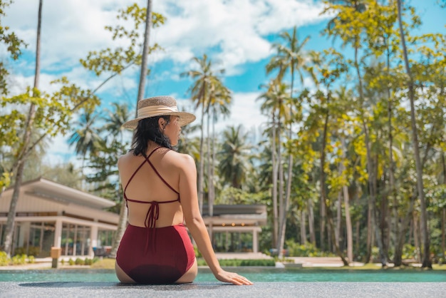 Happy asian woman in red swimsuit and a straw hat relaxing sit\
by the pool at phangnga thailand