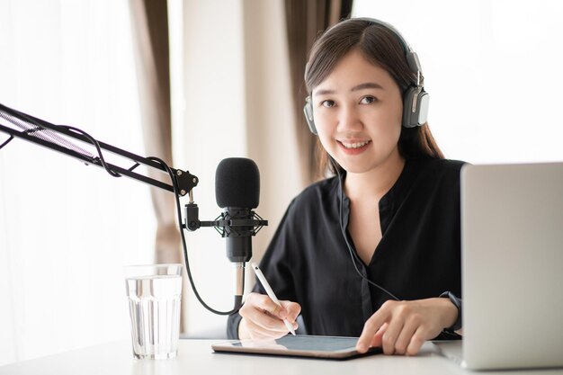 Happy asian woman setting up a living room in her house for\
podcast studio, woman arranging a podcast and online radio station\
at home. professional young podcaster speaking through a\
microphone.