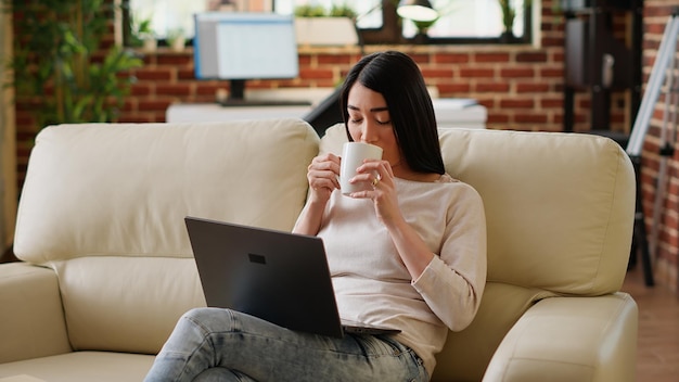 Happy asian woman sipping beverage while working remotely on laptop at home. Smiling heartily young adult person enjoying remote work while sitting on sofa inside modern apartment.