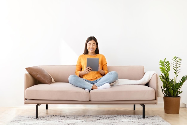 Happy asian woman sitting on couch using tablet at home