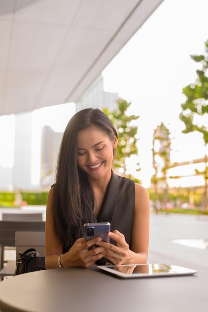 Happy Asian woman sitting outdoors at coffee shop restaurant using mobile phone while smiling