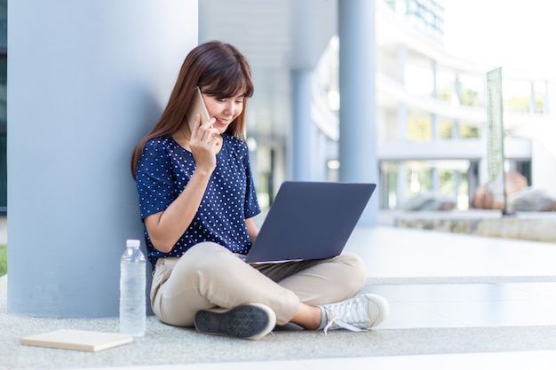 Happy asian woman sitting and talking on phone while using her laptop computer outside of her office