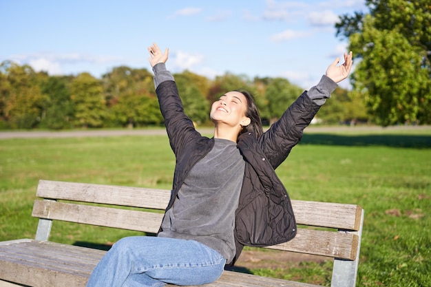 Happy asian woman stretching her hands sitting on bench with excited face smiling pleased feeling fr