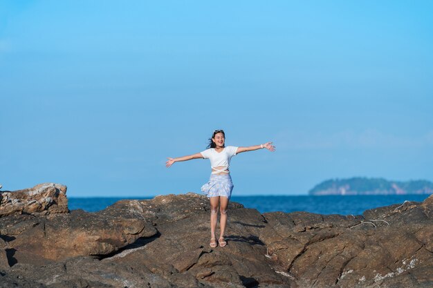 푸른 바다 근처 바위에 서 있는 수영복을 입은 행복한 아시아 여성, 팔을 벌리고 푸른 하늘이 있는 바위 해변에서 휴가, 자유 자연 여행 개념