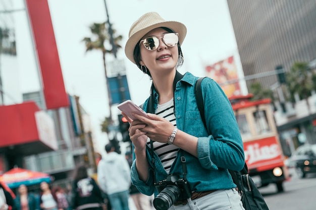 Happy asian woman tourist smiling and walking in street using smartphone and looking around finding direction on online map. blurred view in back with red bus on road in usa lifestyle hollywood.