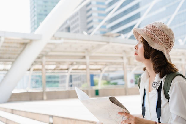 Happy Asian woman traveler tourist wanderer with trendy look searching direction on location map while traveling abroad in summer