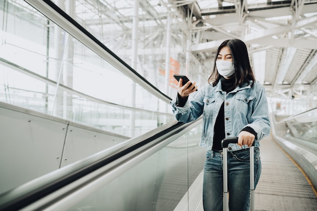 에스컬레이터에 서있는 스마트 폰을 사용하여 마스크를 쓰고 행복 한 아시아 여자 여행자