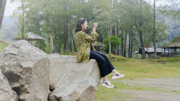 Photo happy asian woman traveller sitting on rock and take the nature picture