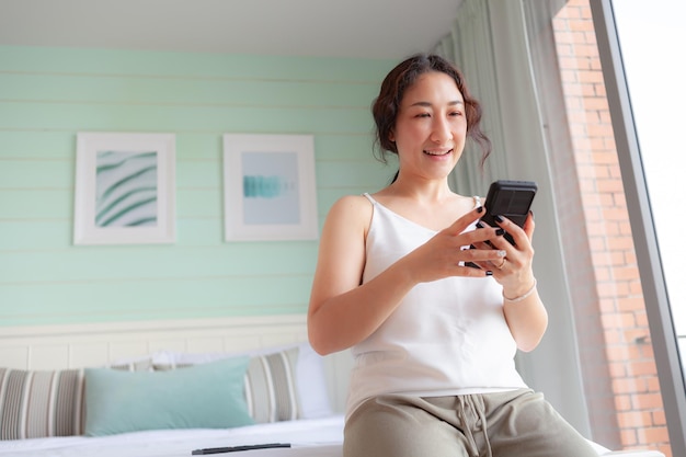 Счастливая азиатская женщина, использующая мобильный телефон дома Азиатская домохозяйка проверяет социальные сети, держит смартфон, расслабляется