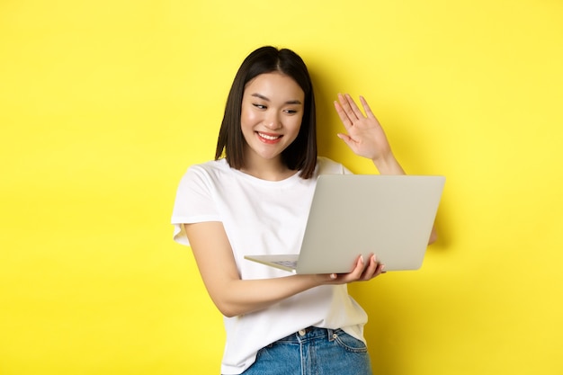 노트북에 행복 한 아시아 여자 화상 채팅, 컴퓨터 카메라에 손을 포기 하 고 인사, 노란색 위에 서.