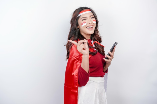 A happy Asian woman wearing headband holding her phone and pointing copy space beside her isolated by white background Indonesia39s independence day