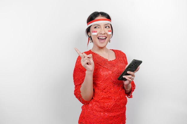 A happy asian woman wearing red kebaya and headband holding her phone and pointing copy space on top of her isolated by white background indonesia's independence day