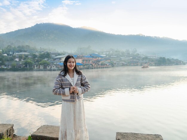 하얀 드레스와 겨울 재킷을 입은 행복한 아시아 여성은 태국의 매홍손(Mae Hong Son)에 있는 반락 타이(Ban Rak Thai) 마을에서 아침에 호수 전망에 안개가 낀 호숫가에 서 있습니다.