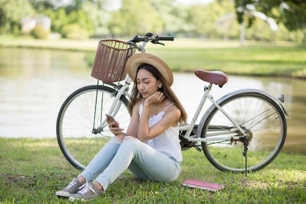 Happy asian woman with hat play smartphone on grass field near bicycle and lake at urban park