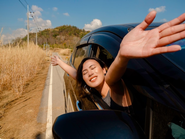 사진 짧은 머리를 한 행복한 아시아 여성이 눈을 감고 손을 들고 차를 타고 여행하는 동안 자유를 느끼고 있습니다. 매력적인 여성 여행자들은 도로 위의 차 밖 풍경을 보며 웃고 있습니다.