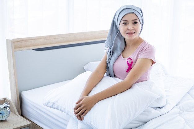 머리 스카프를 두른 분홍색 리본을 쓴 행복한 아시아 여성 유방암 환자는 화학 요법 치료 후 침대에 앉아 집, 건강 관리, 의학 개념의 침실에 앉아 있습니다.