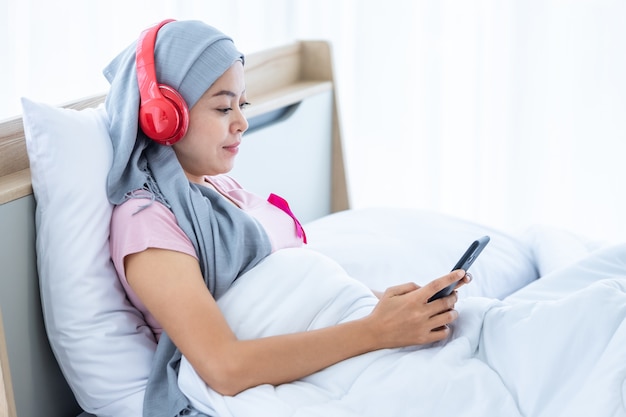 Happy a asian women disease mammary cancer patient with pink ribbon wearing headscarf in headphones is listening to music with smartphone After treatment to chemotherapy sit on bed In the bedroom