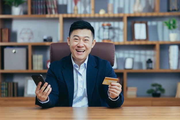 Happy Asian working in the office holding a bank credit card and phone for online shopping looking at the camera and smiling