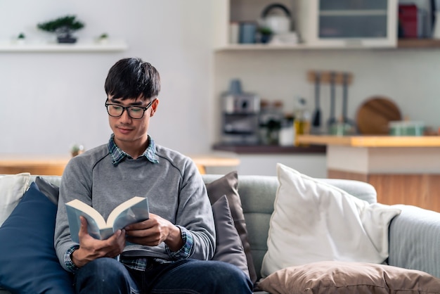 Happy asian young adult man sitting on sofa at living room reading fiction book.