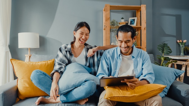 Happy asian young attractive couple man and woman sit on couch use tablet for shopping online furniture in new house