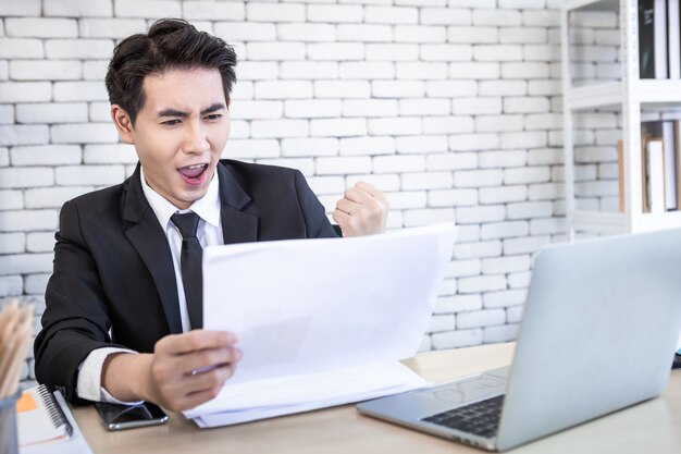 Happy of asian young businessman see a successful business plan on the laptop computer and pen on wooden table background in office,business expressed confidence embolden