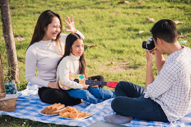 사진 행복한 아시아계 젊은 가족 아버지, 어머니, 아이가 화창한 날 정원 공원에서 피크닉을 즐기며 복고 카메라로 사진을 찍으며 풀밭 파티에 앉아 야외에서 즐거운 시간을 보냅니다.