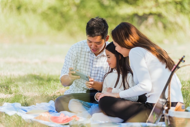 행복한 아시아의 젊은 가족 아버지, 어머니, 어린 소녀가 여름 정원 공원에서 모바일 스마트폰으로 셀카를 찍은 후 피크닉 담요 루킹 사진에 앉아 야외에서 즐거운 시간을 보내고 있습니다.