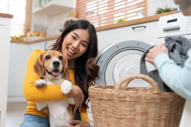 洗濯機に服を入れながらビーグル犬を抱きしめてカメラを見る幸せなアジアの若い女性