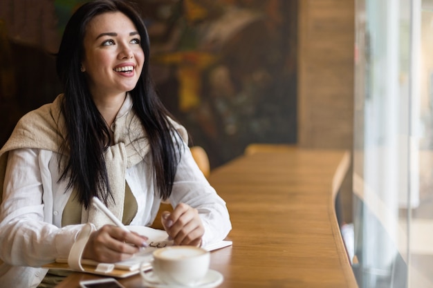 Счастливая азиатская молодая женщина, сидящая в кафе с кофейной чашкой, делая заметки, наслаждаясь перерывом