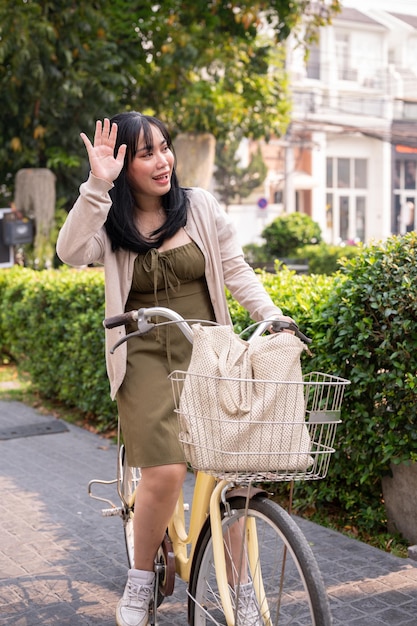 A happy Asian young woman waves her hand to greet her friend while riding a bicycle in the city