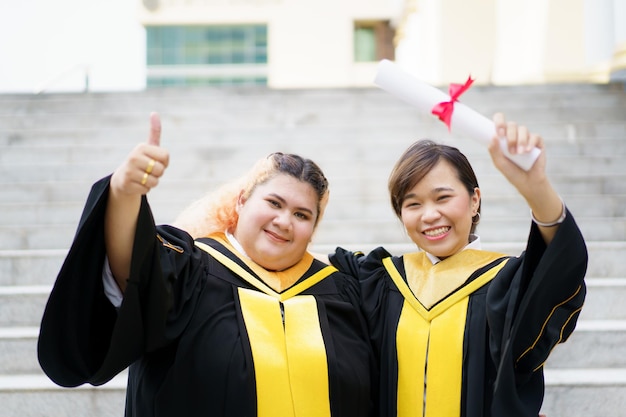 Happy asian young women in the master degree gown showing a
diploma in their hand close up portrait of confident college
students in gown in the graduation ceremony