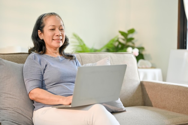 幸せなアジアの退職した女性は、居間でくつろぎながらラップトップで映画を見て楽しんでいます