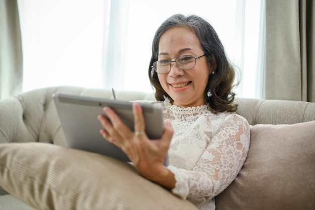 Happy Asianaged woman in eyeglasses sits on sofa in living room using digital tablet