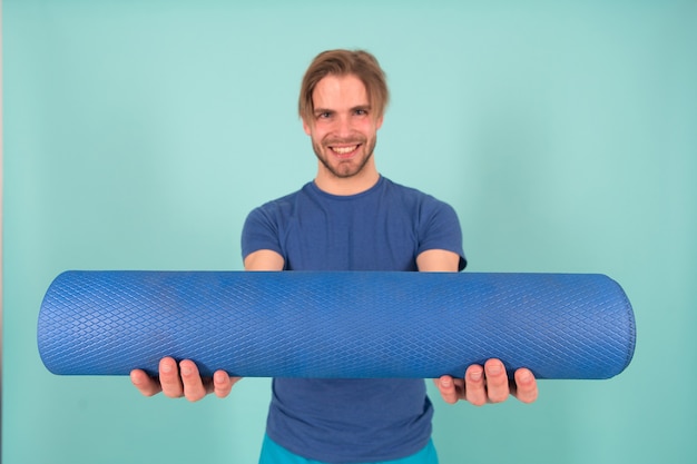 Happy athlete smile with yoga mat. Fashion sportsman in blue tshirt and shorts. Man with stylish bristle and hair. Gym equipment for training and workout. Yoga or pilates and sport activity.