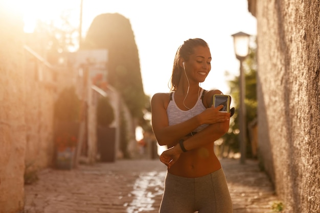 Happy athletic woman adjusting smart phone on her arm band while preparing for exercise in the city