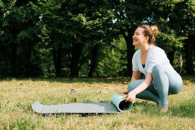 자연에서 맨발로 잔디에 앉아있는 동안 요가 매트를 비틀는 행복한 운동 여자 웃는 운동가의 측면보기 자연에서 훈련의 끝 또는 시작
