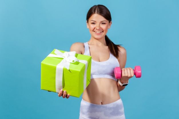 Happy athletic woman in white sportswear holding green gift box and pink dumbbell