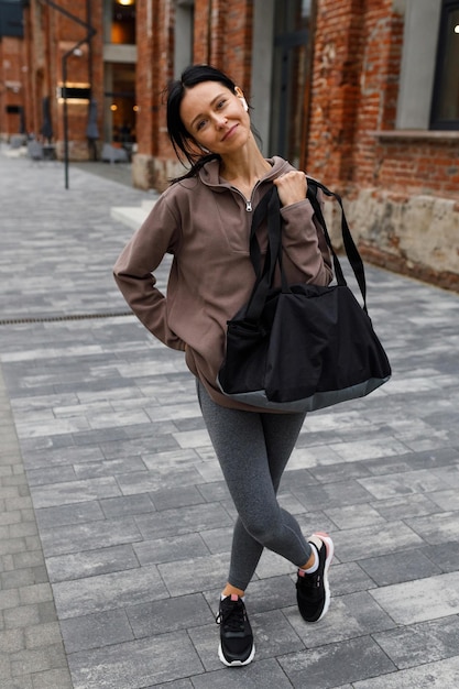 Happy athletic woman with sports bag posing standing near brick building