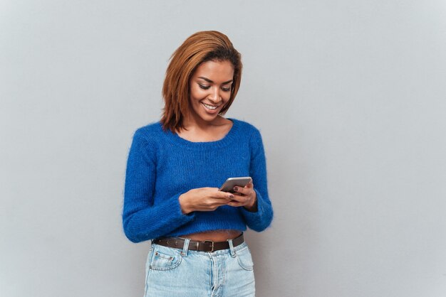 Happy attractive african woman in sweater and jeans writing message on phone in studio. Isolated gray background